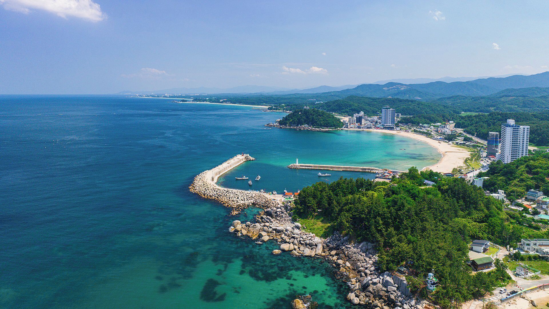 A coastal area with a rocky breakwater, clear blue waters, and a mix of natural and urban landscapes.