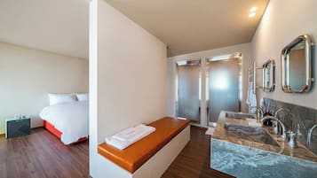 Habitación de hotel con cama, una banca, ducha y baño con lavamanos y espejo.