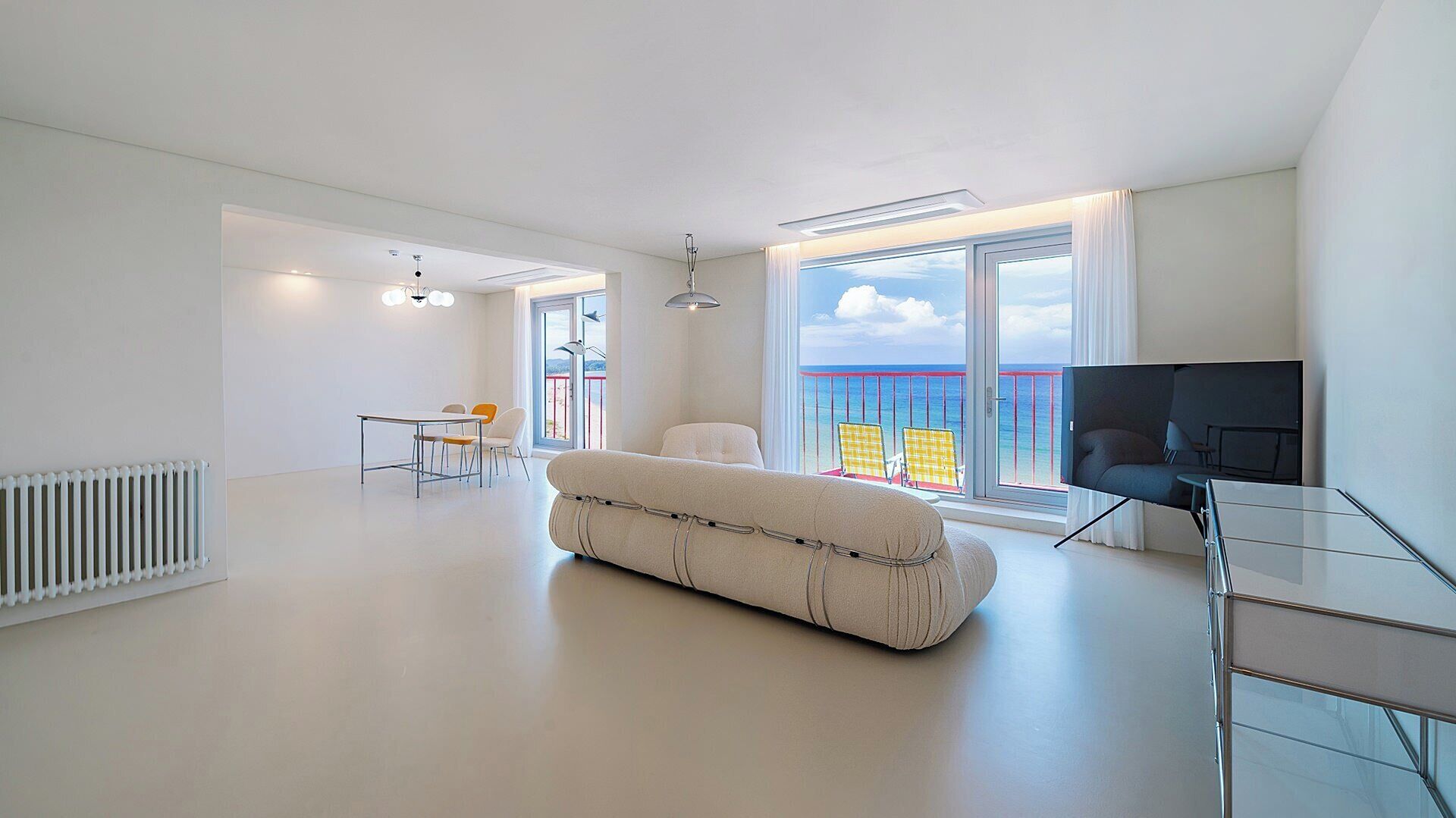 A modern living room with a sofa, a TV, and a balcony with a view of the sea.