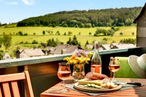 A table with a bottle, glasses, flowers, cheese, and bread on a balcony overlooking a green landscape.