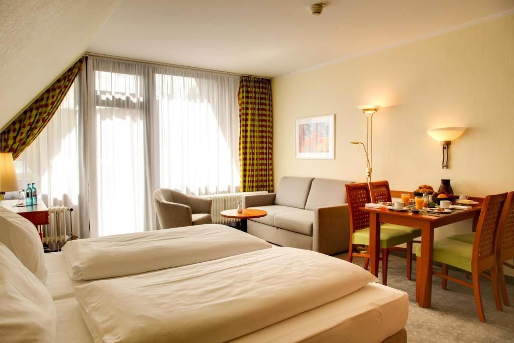 A hotel room with a bed, a sofa, a dining table with chairs, a lamp, and a window with curtains.