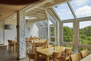 A dining area with wooden tables and chairs, a stone wall, and large windows offering a view of greenery.
