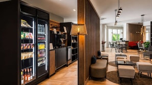 Um hall de hotel moderno com uma geladeira abastecida com bebidas, um painel de parede em madeira e um espaço de estar com mesas e cadeiras.