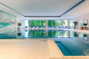Ein Innenpool mit Blick auf Bäume durch große Fenster, blaue Liegestühle und einem Wandgemälde.