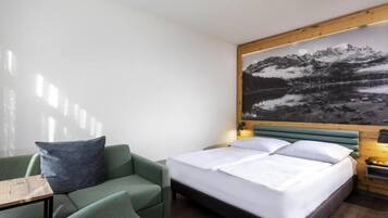 A hotel room with a bed, a green armchair, a wooden headboard, and a wall-mounted picture of a mountain landscape.