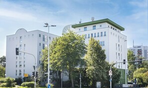 Un bâtiment moderne à la façade blanche et au toit végétalisé, entouré d’arbres et d’éléments urbains.
