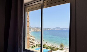 Blick aus dem Fenster auf eine Küstenlandschaft mit Strand, Pool und einer Stadt im Hintergrund.