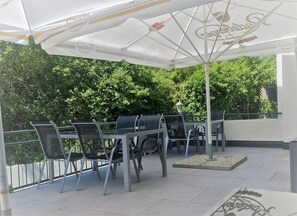 Een terras buiten met metalen stoelen en een tafel onder een witte parasol.