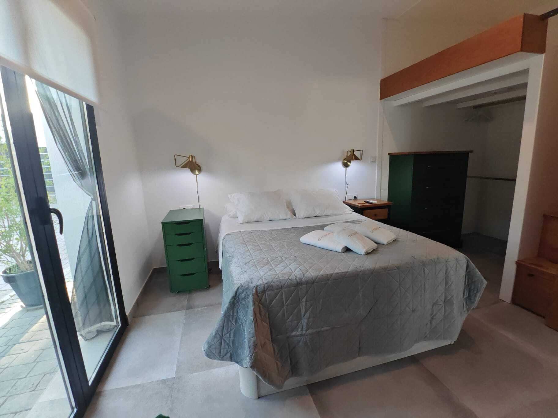 Een slaapkamer met een bed, twee bedlampen, een groen opbergmeubel en uitzicht door een schuifdeur.