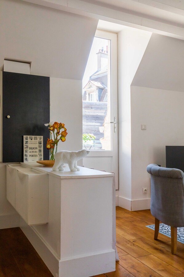 Una habitación con una consola blanca, una silla blanca, una ventana con vistas a edificios y un cuadro en la pared.