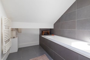 Um banheiro moderno com pia branca, paredes revestidas de azulejos escuros, radiador branco e um banquinho de madeira.
