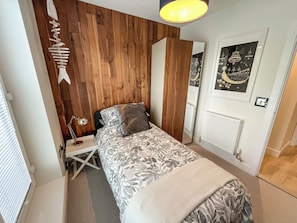 A bedroom with a wooden wall, a bed with patterned bedding, a bedside table with a lamp, and a mirror. - Whitstable May House (Whitstable)