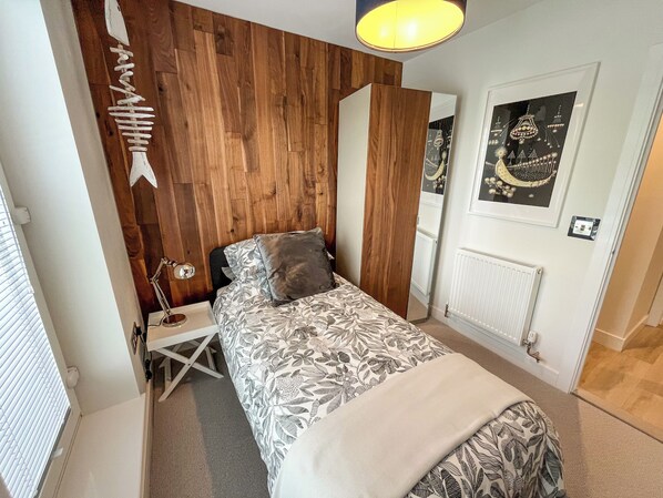 A bedroom with a wooden wall, a bed with patterned bedding, a bedside table with a lamp, and a mirror. - Whitstable May House (Whitstable)