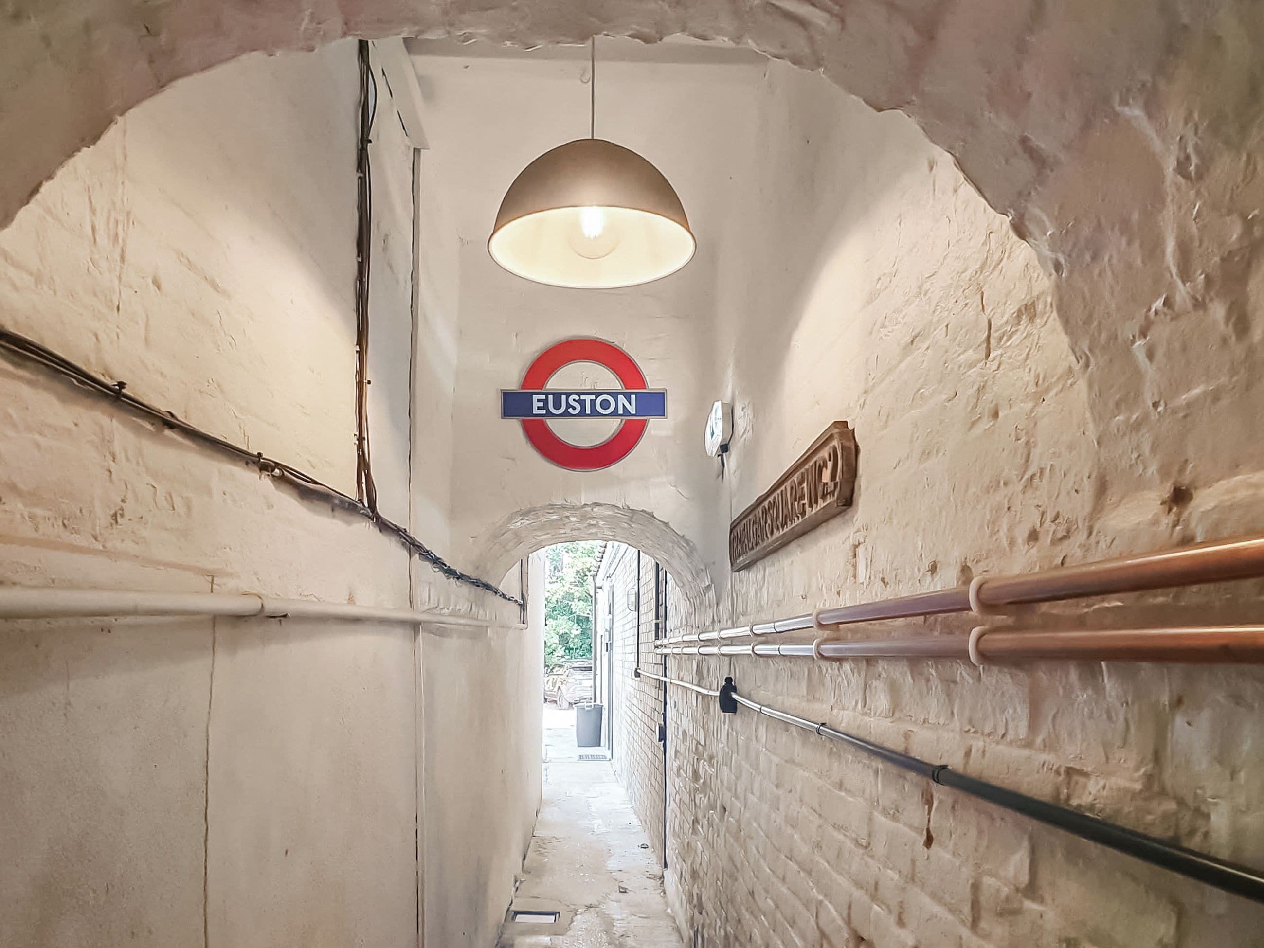 A tunnel-like passageway with a circular sign indicating 'Euston' station, a hanging light fixture, and a metal handrail on the right.