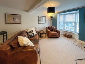 A living room with a brown leather sofa, a small armchair, a floor lamp, and a large window with blinds.