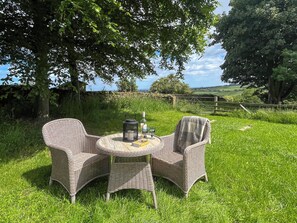 Une table en osier avec deux chaises, une bouteille et un verre, le tout placé dans un cadre extérieur verdoyant.