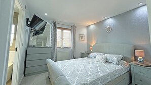 Un dormitorio con cama, mesitas de noche, televisor montado en la pared, ventana con cortinas y una decoración en forma de corazón.
