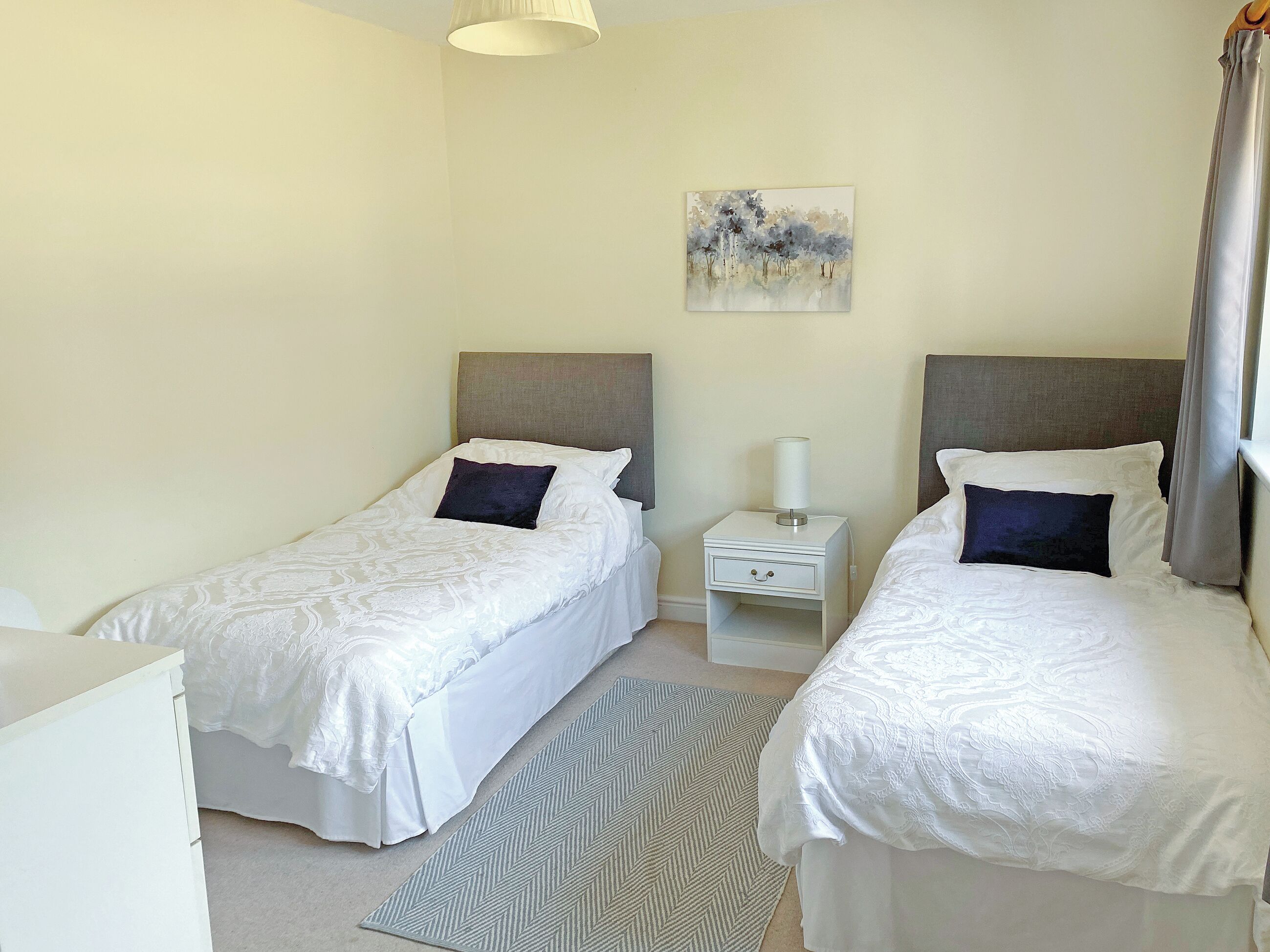 A hotel room with two single beds, a nightstand with a lamp, a painting on the wall, and a herringbone-patterned rug.