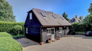 A small, dark wooden house with a sloped roof, a gravel driveway, and a well-maintained garden.