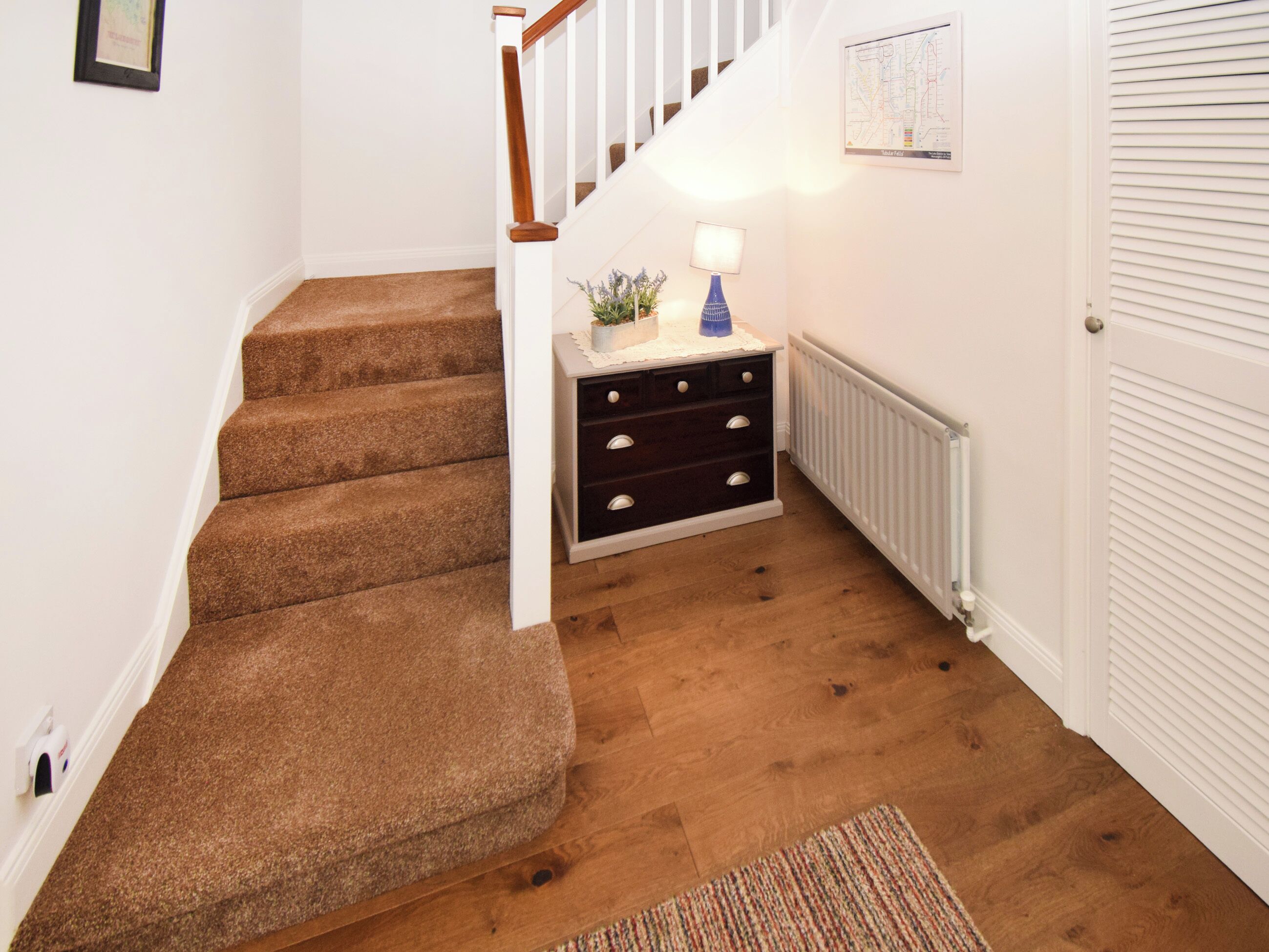 Eine Treppe mit Teppichstufen, ein kleiner Nachttisch mit Lampe, eine Heizung und eine Tür mit Lamellen.