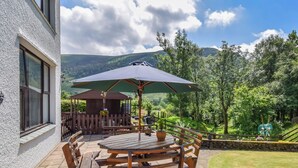 A patio with a wooden table and chairs, a green umbrella, and a view of a lush garden and hills. - 2 bedroom accommodation in Ennderdale (Ennderdale)