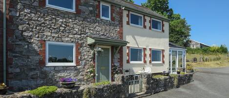 Une maison en pierre à deux étages, dotée d’un mur en pierre, d’une porte verte et d’un petit porche avec une chaise blanche.