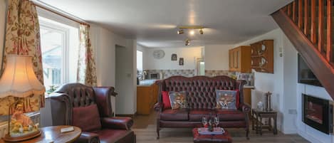 Una sala de estar con sofá, sillón y reposapiés, una chimenea y una cocina al fondo.