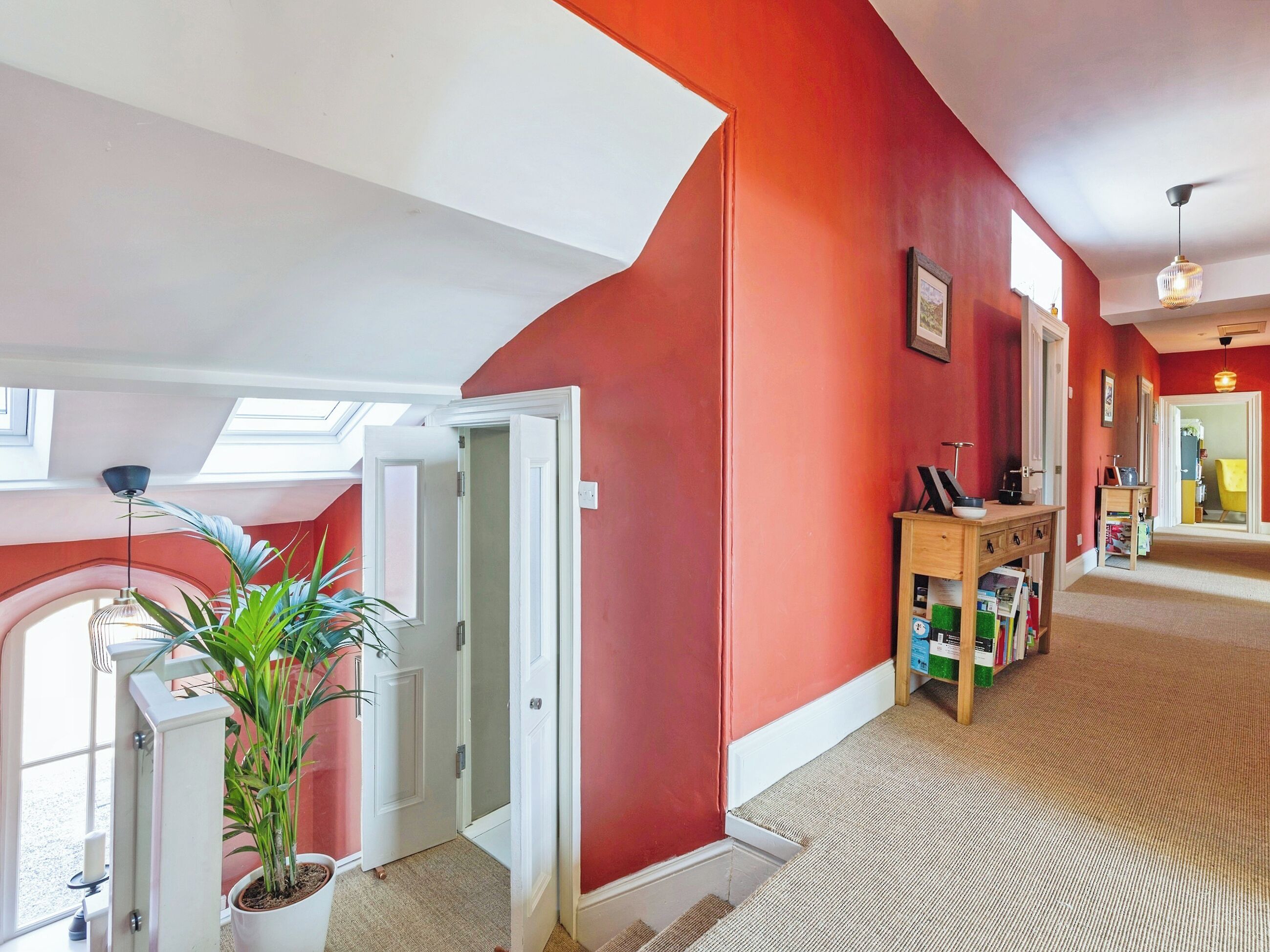 A hallway with a red wall, a white door, a plant, and a wooden desk with books.