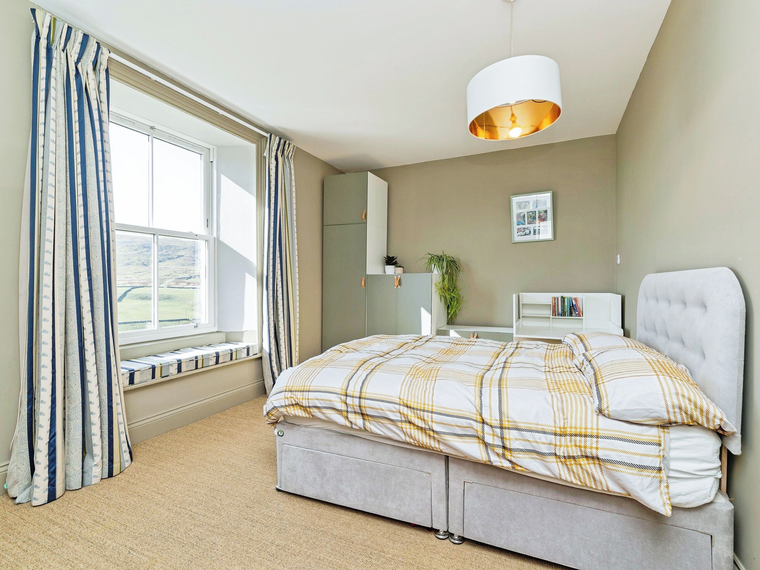 A bedroom with a bed, a window with curtains, a bedside table, a lamp, and a wall-mounted picture.