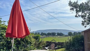 En terrasse med et bord og stole, en rød parasol og udsigt over et grønt område og bakker i det fjerne.