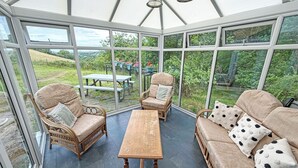 A conservatory with wicker furniture, a wooden table, and a view of the garden. - Hendre Aled Farmhouse (Llanssannan)