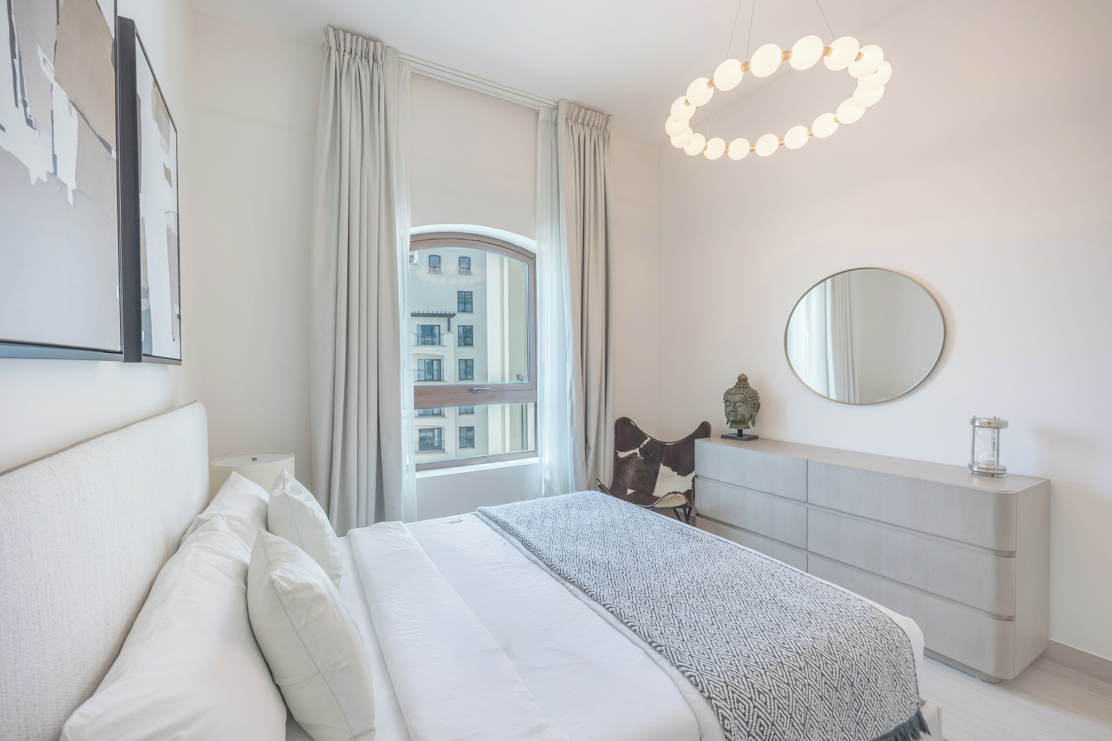 A modern bedroom with a bed, a dresser, a mirror, and a view of buildings outside the window.