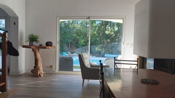 Una habitación amplia con una escalera de madera, un gran ventanal corredero que da a una piscina, una mesa de madera y una silla.