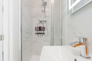 A modern bathroom with a glass shower enclosure, a white sink with a chrome faucet, and a marble wall. - Newly Renovated 4BDR Home Near Notting Hill (Greater London)
