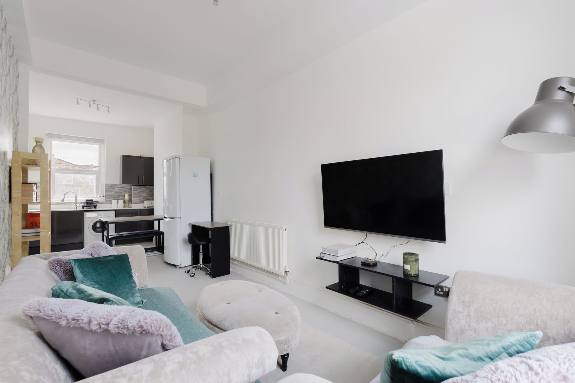 Un salon moderne avec un téléviseur à écran plat, un canapé, des poufs et une cuisine en arrière-plan.