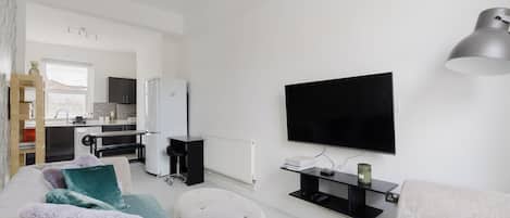 A modern living room with a flat-screen TV, a sofa, ottomans, and a kitchen in the background.