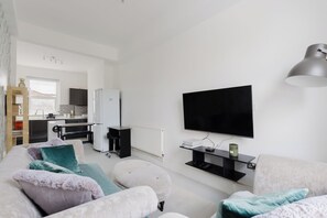 A modern living room with a flat-screen TV, a sofa, ottomans, and a kitchen in the background. - Newly Renovated 4BDR Home Near Notting Hill (Greater London)