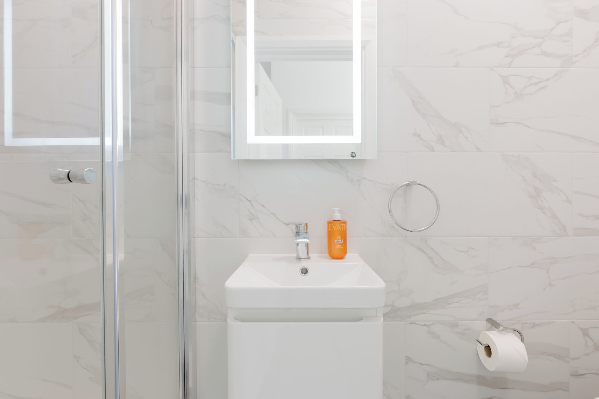 Een badkamer met een douchecabine met glazen deur, een witte wastafel met zeepdispenser, en een toiletrolhouder.