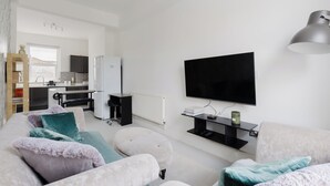 A modern living room with a flat-screen TV, a sofa, ottomans, and a kitchen in the background. - Newly Renovated 4BDR Home Near Notting Hill (Greater London)