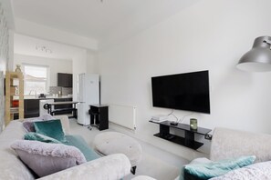 Ein modernes Wohnzimmer mit Fernseher, einer Couch, Poufs und einer Küche im Hintergrund.