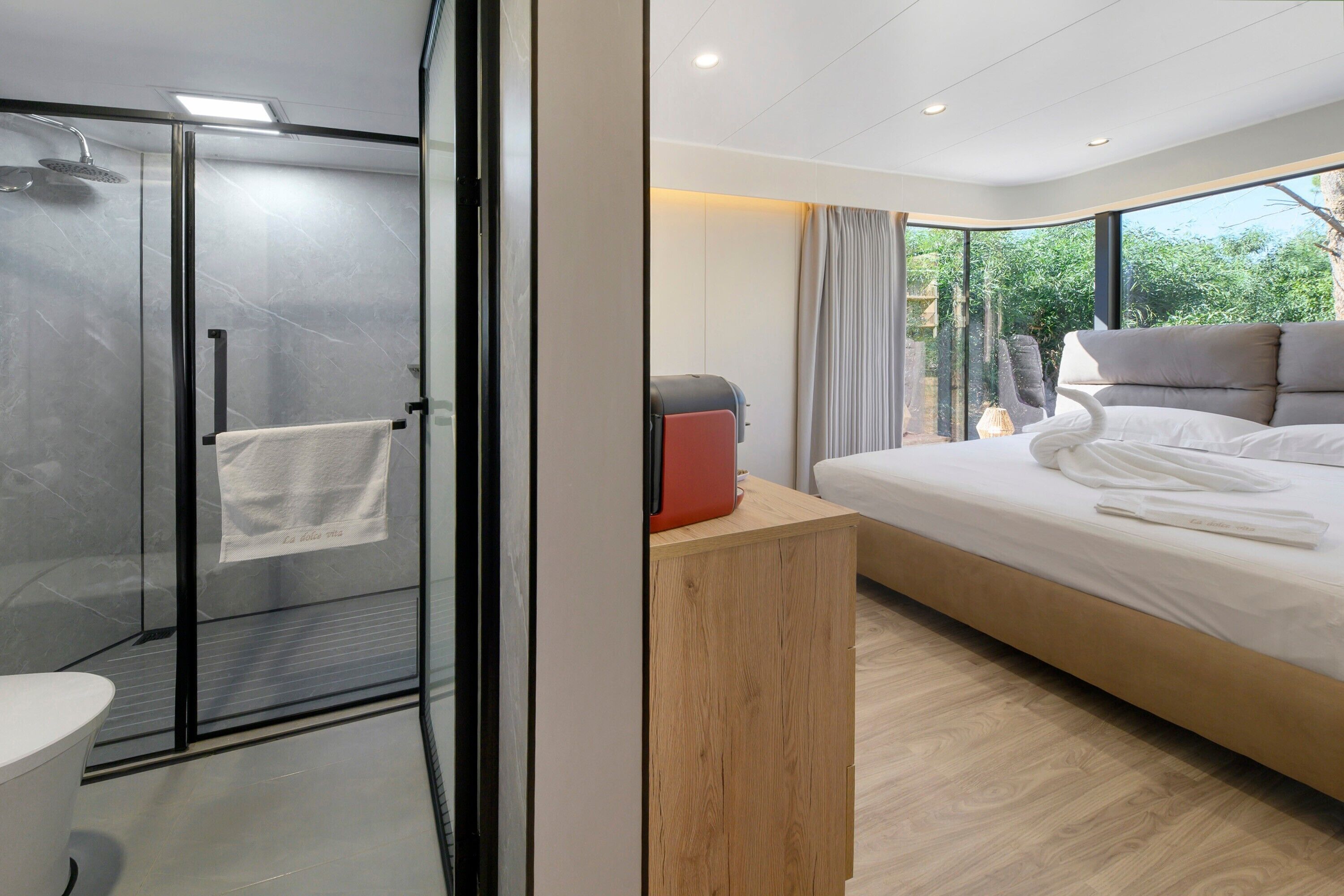 A modern hotel room with a glass-enclosed shower, a wooden bedside table, a bed with white linens, and a view of greenery outside.