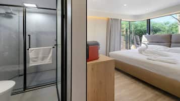 A modern hotel room with a glass-enclosed shower, a wooden bedside table, a bed with white linens, and a view of greenery outside.