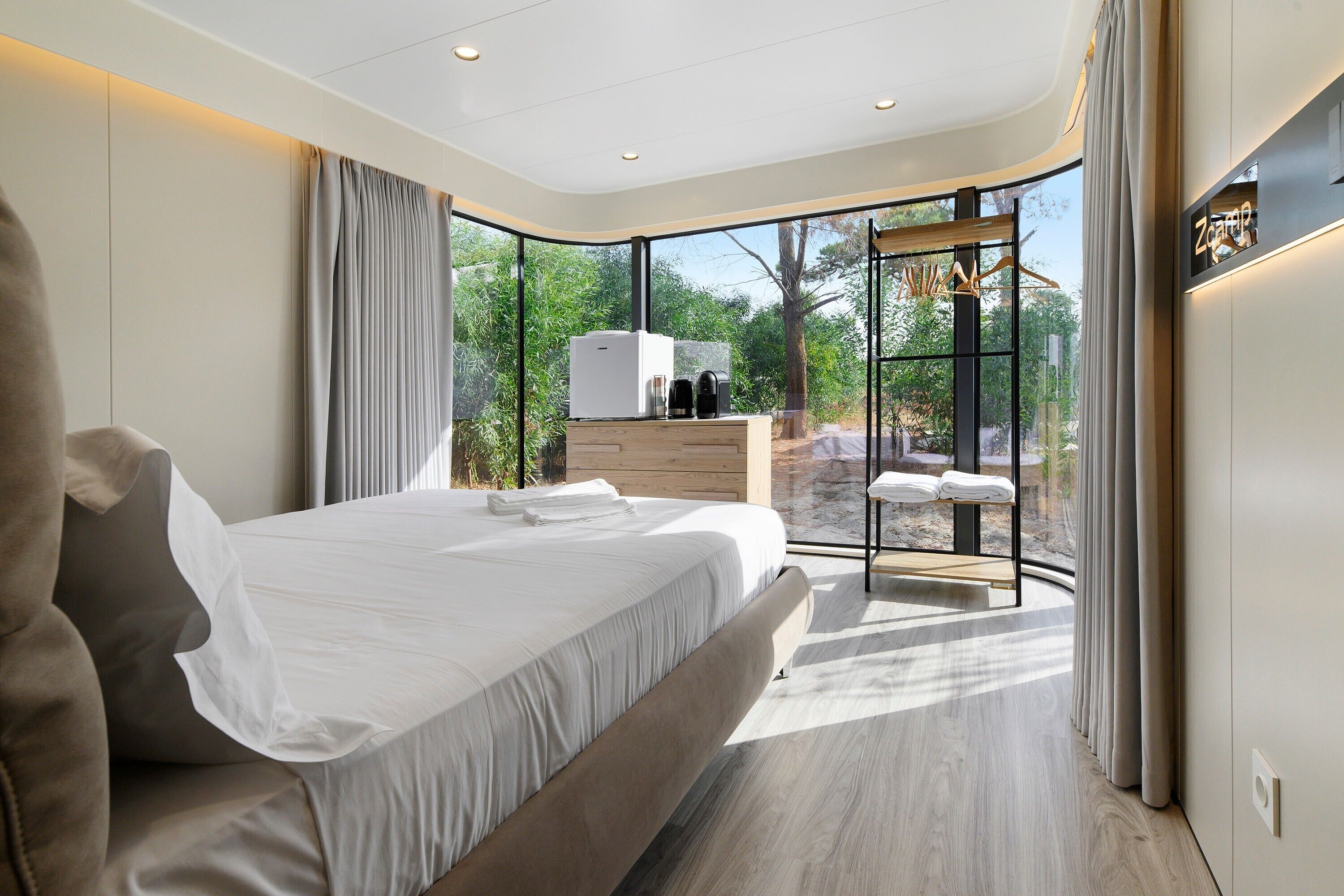 A modern bedroom with a large bed, a wooden dresser, a seating area with a table, and a view of greenery through large windows.