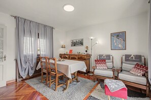 A dining area with a wooden table, chairs, and a rug. There is a window with curtains, a lamp, and a framed picture on the wall.