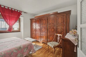 Une chambre à coucher avec une armoire en bois, un lit recouvert d’une couvre-lit à motifs, une chaise et une petite table agrémentée d’un objet décoratif.