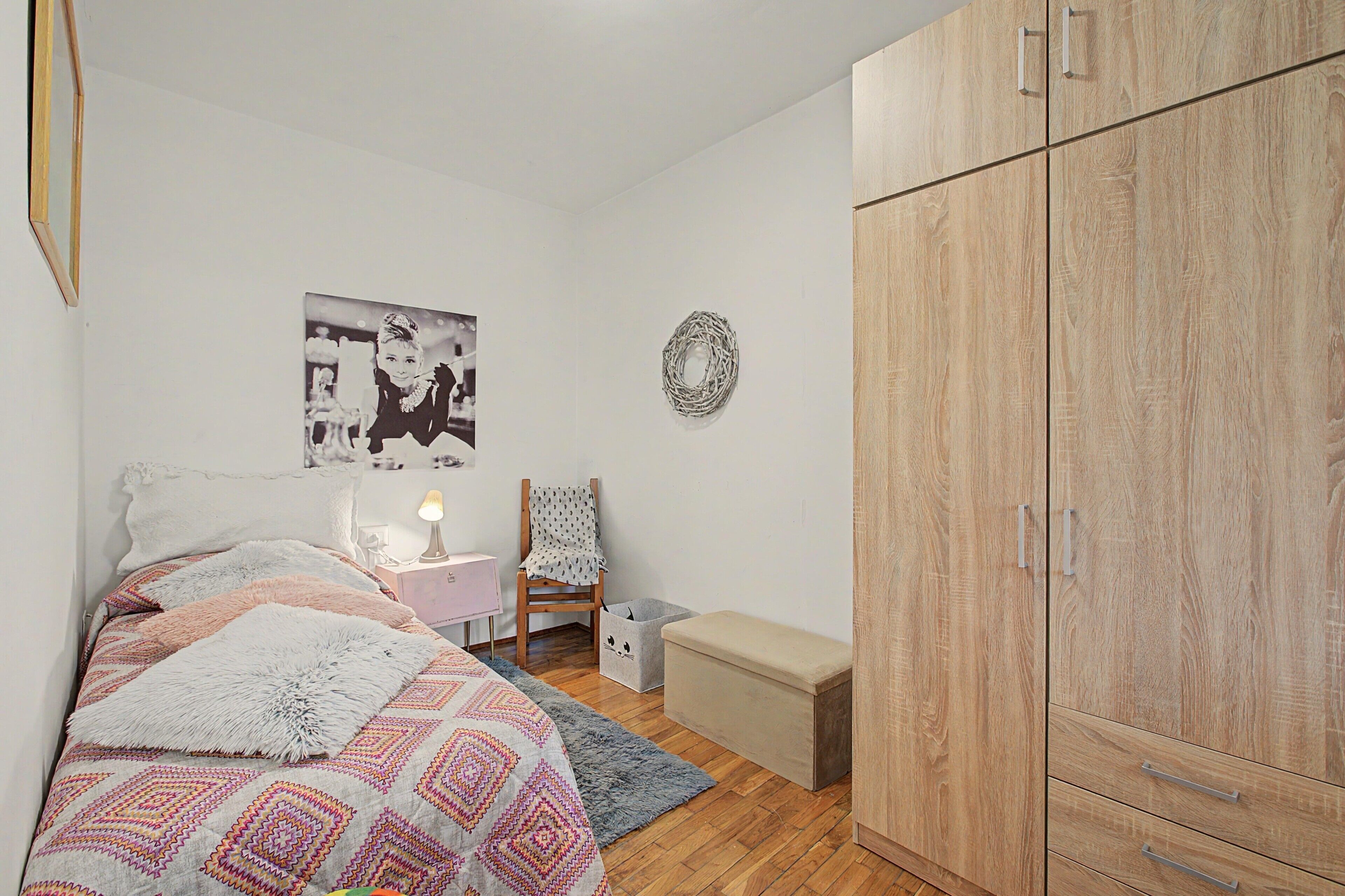 Un dormitorio con cama, una silla, una mesita, un armario y una pared con un cuadro.