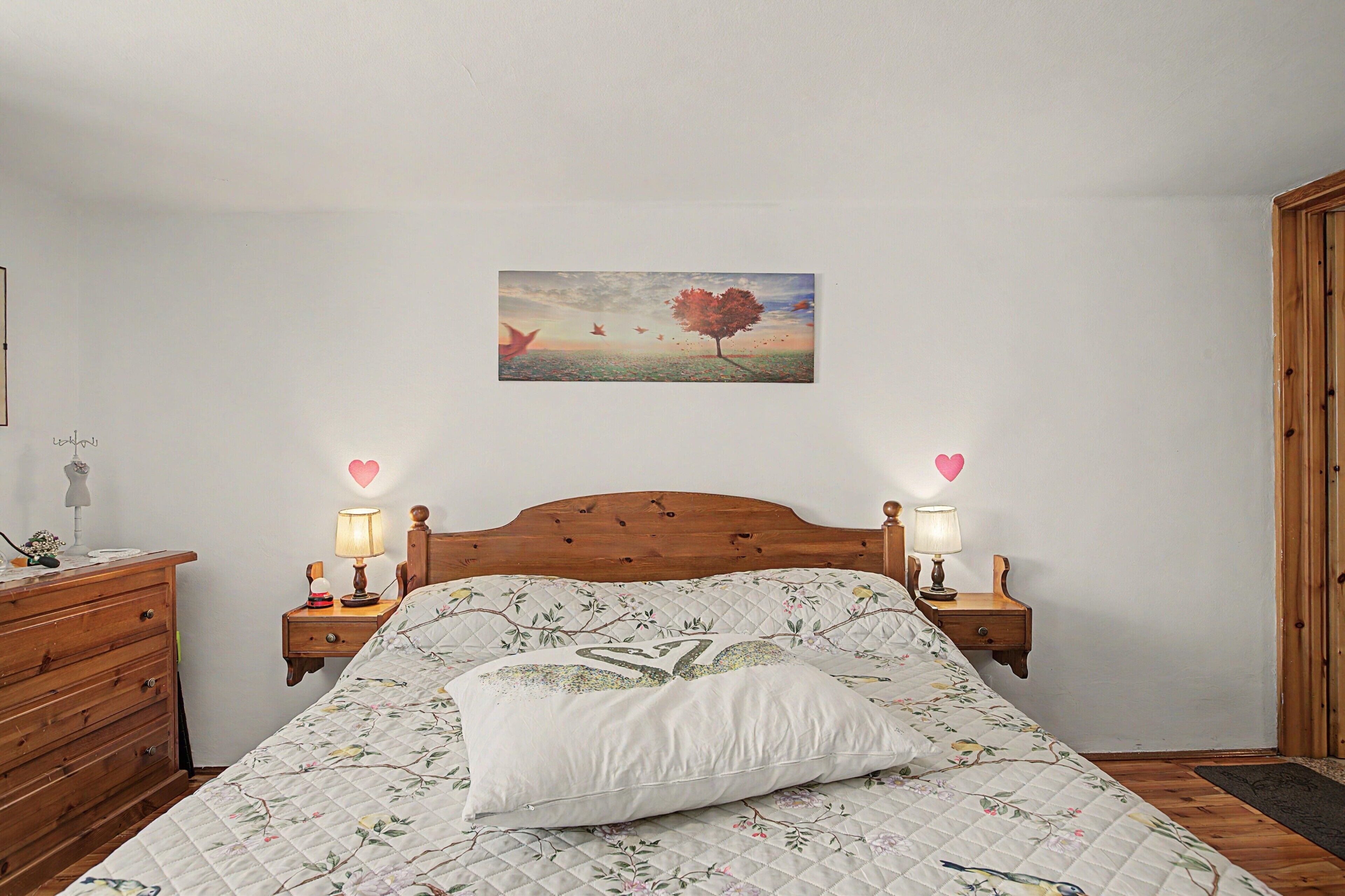 Une chambre avec une tête de lit en bois, une couvre-lit matelassé à motifs floraux, deux lampes de chevet, une table de chevet avec une lampe, une commode et un tableau accroché au mur représentant un arbre et des oiseaux.
