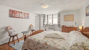 Un dormitorio con cama, una silla, tocador, radiador y ventana con cortinas.
