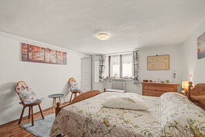 Une chambre à coucher avec un lit, une chaise, une commode, un radiateur et une fenêtre avec des rideaux.