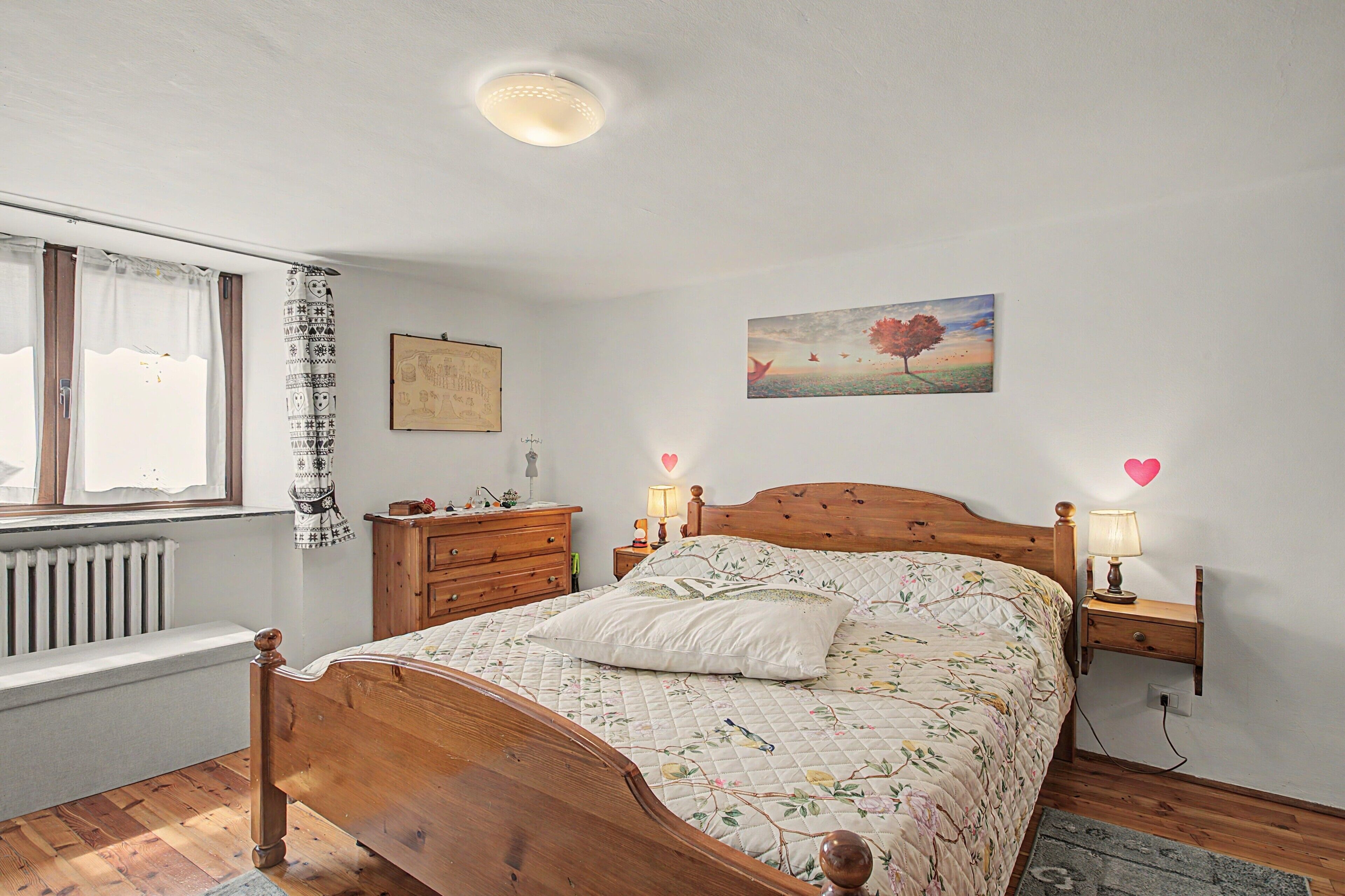 Un dormitorio con una cama de madera, una mesita de noche con lámpara, un aparador, una ventana con cortinas y un cuadro en la pared.